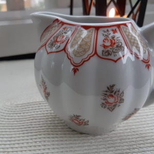 Può includere: Una lattiera in ceramica bianca con corpo arrotondato e manico. La lattiera è decorata con un motivo floreale rosso e oro sul bordo e motivi floreali più piccoli sul corpo. La lattiera è appoggiata su una superficie bianca strutturata.