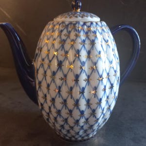 May include: A white porcelain teapot with a blue and gold pattern. The teapot has a rounded body, a curved spout, and a handle. The lid is topped with a small knob. The pattern is a blue lattice with gold accents.