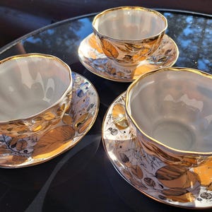 Peut inclure: Trois tasses à thé en porcelaine blanche avec soucoupes assorties, dotées d'un bord festonné et d'un motif botanique doré métallique. Le design présente des feuilles et des branches dorées détaillées sur un fond blanc avec des bords dorés.