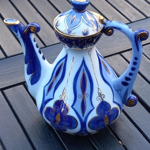Könnte beinhalten: Dekorative Porzellan-Teekanne mit Deckel, mit blau-weißem Blumendesign mit goldenen Akzenten. Die Teekanne hat einen gebogenen Griff und Ausguss, und der Deckel ist mit einem goldenen Knopf versehen. Die Teekanne ist etwa 20 cm hoch.