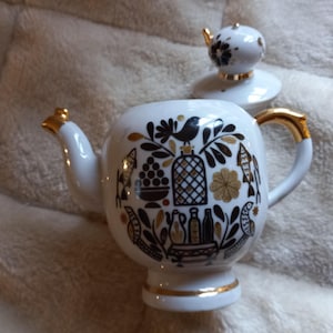 以下が含まれることがあります： 黒と金の装飾模様が施された、蓋付きの白いセラミックティーポット。 ティーポットは、金色のアクセントが付いた湾曲した注ぎ口とハンドルを備えています。 蓋は別になっており、お揃いのデザインが施されています。 柔らかい白い表面にあります。