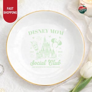 a white plate with a green and gold design, which includes a castle and a coffee cup, along with the text "Disney Mom Social Club" and "This mom runs on coffee and magic".