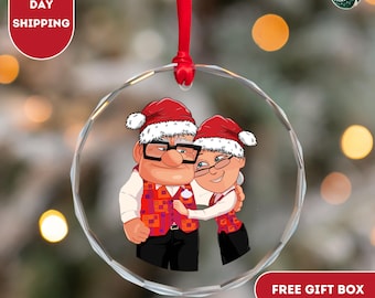 Könnte beinhalten: Ein transparenter Weihnachtsbaumschmuck aus Glas mit einem Cartoon-Paar, das Weihnachtsmützen und rot-weiße Westen trägt. Das Paar umarmt sich und lächelt. Der Schmuck hat ein silbernes Band, das oben befestigt ist.