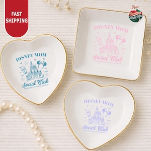 three decorative dishes with Disney-themed designs, including a heart-shaped dish, a square dish, and a smaller heart-shaped dish. The dishes are placed on a white surface, and there is a string of pearls nearby.