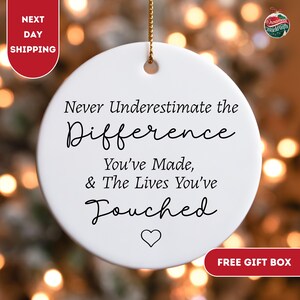 May include: A white ceramic ornament with gold string for hanging. The ornament has the text "Never Underestimate the Difference You've Made, & The Lives You've Touched" in black script. A heart symbol is at the bottom.
