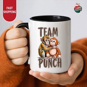 A person is holding a white mug with a cartoon image of two monkeys hugging each other.