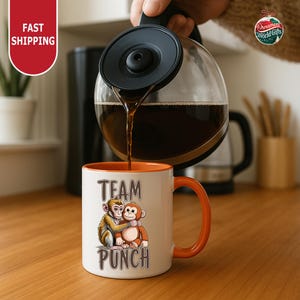 A person is pouring coffee from a coffee pot into a white mug with an orange handle, which has a cartoon monkey design and the text "TEAM PUNCH" printed on it.