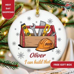 May include: A white ceramic Christmas ornament featuring a toolbox design with tools, an orange hard hat with "2025", and the text "Oliver I can build that". The ornament has a gold string for hanging and snowflake accents.
