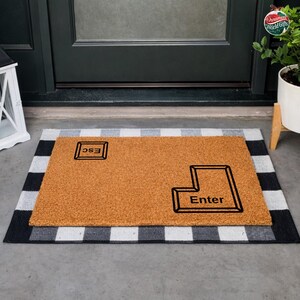May include: A rectangular doormat made of brown coir with the word "Enter" and "ESC" in black. The doormat is placed on a black and white checkered rug, in front of a black door.
