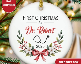 May include: A white ceramic Christmas ornament with the text "FIRST CHRISTMAS AS Dr. Robert 2025". The ornament features a stethoscope graphic, a red bow, and floral accents. The ornament has a gold string for hanging.