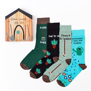 Puede incluir: Cuatro pares de calcetines coloridos con juegos de palabras sobre el tema de la jardinería. Los calcetines son de color verde, negro, gris y azul. Los calcetines están empaquetados en una caja que parece un cobertizo con las palabras "Urban Eccentric Sock Shed" en la parte delantera.
