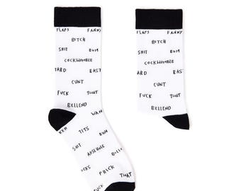 May include: A pair of white socks with black heels and toes. The socks are covered in various black text, including words like "FLAPS", "FANNY", "BITCH", "SHIT", "FUCK", and others. The socks are displayed against a white background.