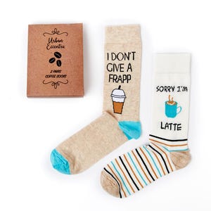 May include: Two pairs of cotton socks with coffee-themed designs. One pair features a beige sock with a brown and white illustration of a coffee drink and the text "I don't give a frapp". The other pair features a white sock with brown and white stripes and a brown and white illustration of a coffee mug and the text "Sorry I'm latte".