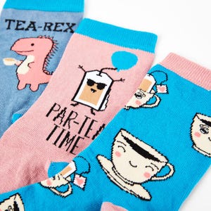 May include: Three pairs of colorful socks with fun tea-themed designs. One pair features a pink dinosaur with the text "Tea-Rex", another pair has a tea bag with sunglasses and the text "Par-Tea Time", and the third pair has a blue background with multiple teacups with faces.
