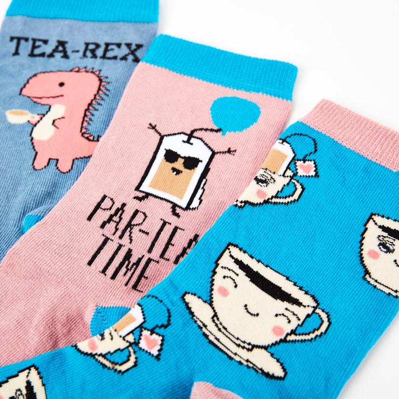 May include: Three pairs of colorful socks with fun tea-themed designs. One pair features a pink dinosaur with the text "Tea-Rex", another pair has a tea bag with sunglasses and the text "Par-Tea Time", and the third pair has a blue background with multiple teacups with faces.