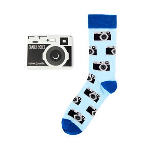 May include: Light blue socks patterned with black and white camera illustrations, featuring a blue cuff and toe. The image also shows a matching box with the text "CAMERA SOCKS" and a camera design.