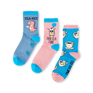 May include: Three pairs of colorful socks with fun tea-themed designs. One pair features a dinosaur with the text "Tea-Rex", another pair has a tea bag with the text "Par-Tea Time", and the third pair has a teacup with the text "Time for Tea".