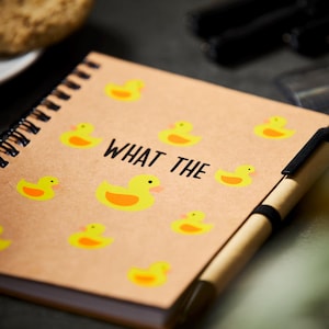 May include: A brown notebook with a spiral binding and a pen attached. The cover has a pattern of yellow rubber ducks and the text "WHAT THE" in black.