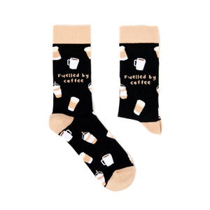 May include: Black socks with a beige heel and toe, patterned with coffee cups and iced coffee drinks. The text "Fuelled by coffee" is printed on the front. A fun accessory for coffee lovers, these socks are a great gift.