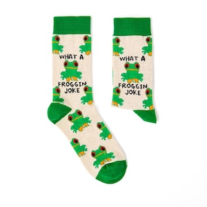 Könnte beinhalten: Ein Paar weiße Socken mit grünem Besatz und einem grünen Froschmuster. Die Socken tragen den Text "What a Froggin Joke".