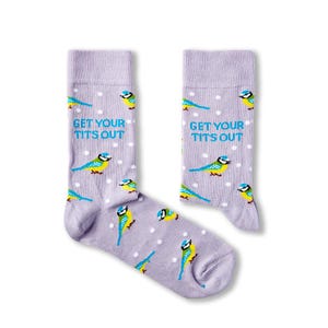 May include: A pair of lavender socks featuring a pattern of blue and yellow birds and white dots. The text "GET YOUR TITS OUT" is printed on the socks.