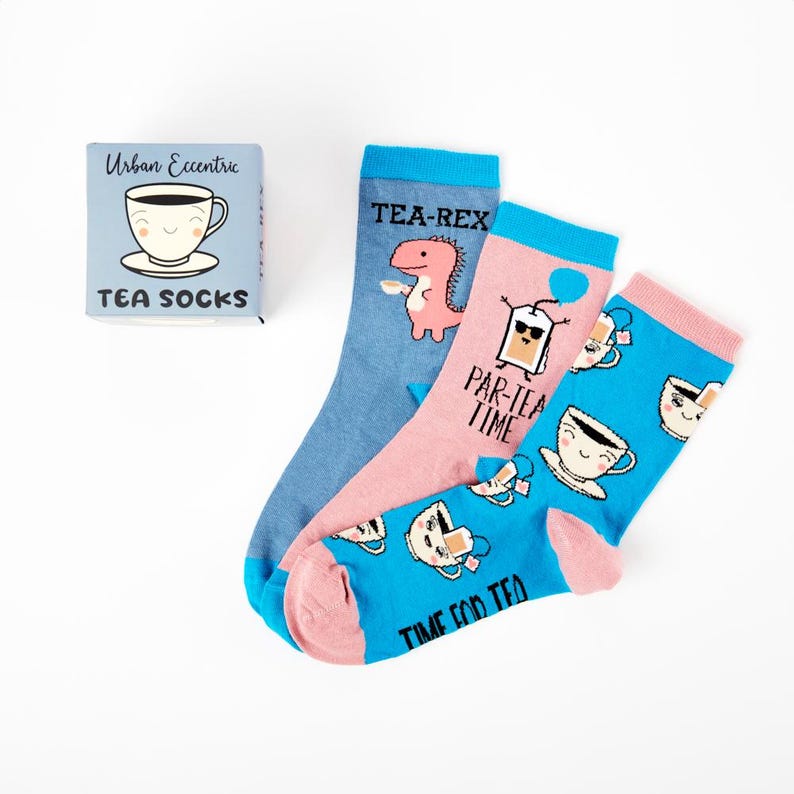 May include: A set of three colorful socks with tea-themed designs. The socks feature a blue sock with a dinosaur holding a teacup, a pink sock with a teabag wearing sunglasses and the text "Par-tea Time", and a blue sock with a teacup with a face and the text "Time for Tea". The socks are packaged in a small blue box with the text "Urban Eccentric Tea Socks".