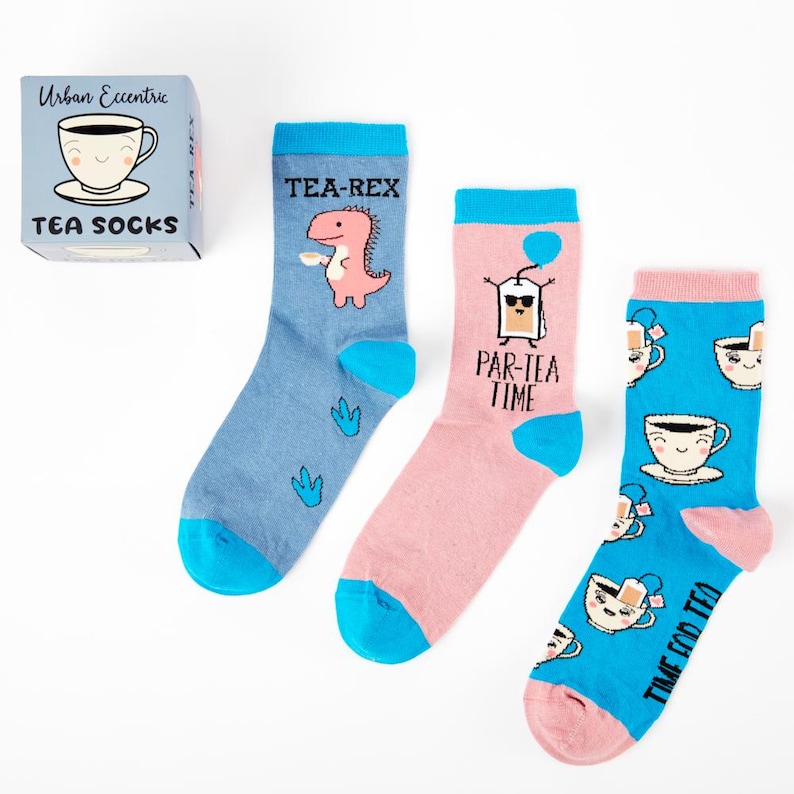 May include: A set of three pairs of colorful socks with tea-themed designs. The socks feature a dinosaur, a tea bag, and a teacup. The socks are packaged in a small box with the words "Tea Socks" on it.