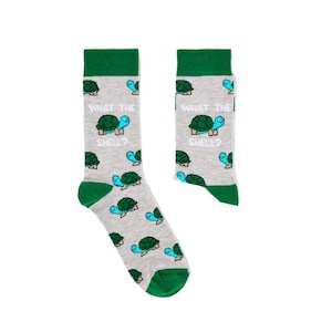 Pode incluir: Um par de meias cinzentas com punhos, calcanhares e dedos verdes. As meias apresentam um padrão repetido de tartarugas de desenho animado e o texto "WHAT THE SHELL?" em branco.