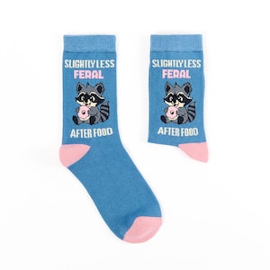 May include: A pair of blue socks featuring a cartoon raccoon with a pink donut. The text "SLIGHTLY LESS FERAL" and "AFTER FOOD" are printed above and below the raccoon. The toe and heel are pink.