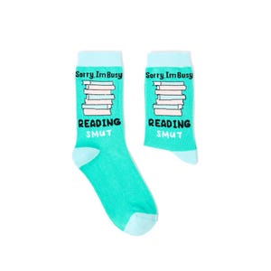 May include: A pair of turquoise socks with light blue details. Each sock has the text "Sorry, I'm Busy" above a stack of books and the words "Reading Smut" below. The socks are set against a white backdrop.