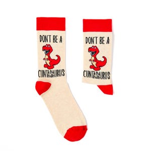 May include: A pair of novelty socks with a red and beige color scheme. The socks feature a red dinosaur graphic and the text "DON'T BE A CUNTASAURUS" in black. The toe and cuff of the socks are red.