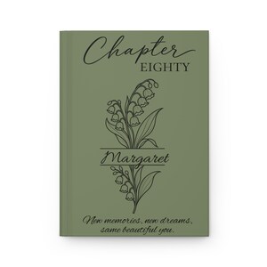 May include: A sage green hardback journal featuring black line art of lily of the valley flowers. The text reads Chapter EIGHTY, the name Margaret, and the phrase New memories, new dreams, same beautiful you. The design is centred.