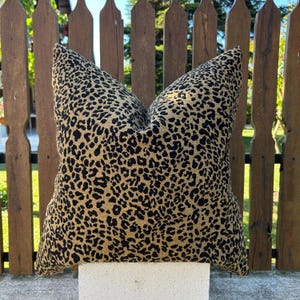 May include: Square cushion with a leopard print pattern. The cushion has a tan background with black spots, creating an animal print design. The cushion is placed on a white surface in front of a wooden fence.