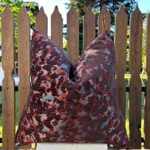 May include: A decorative throw pillow with a textured, abstract pattern in shades of brown, red, and teal. The pillow is square and appears to be made of a woven fabric, adding a touch of color and texture to any living space.