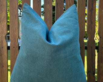 May include: A square teal pillow with a textured appearance. The pillow is made of a soft fabric and is placed in front of a wooden fence. The pillow is a solid color and has a simple, elegant design, suitable for home decor.