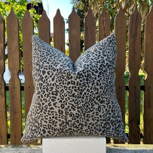 May include: A square cushion featuring a leopard print design in black on a beige background. The cushion is positioned in front of a wooden fence.