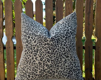 May include: A square decorative pillow with a neutral beige background and a black leopard print pattern. The pillow is placed in front of a wooden fence.