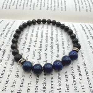 Może przedstawiać: Bransoletka z koralików z połączeniem czarnych koralików z lawy wulkanicznej i ciemnoniebieskich koralików z lapis lazuli. Bransoletka jest nawleczona na elastyczny sznurek i zawiera srebrne metalowe akcenty. Bransoletka jest prezentowana na otwartej książce.