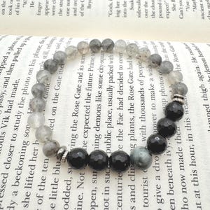 May include: A beaded bracelet featuring a mix of gray and black gemstone beads. The bracelet alternates between faceted black onyx and gray-toned beads, with silver-colored metal accents. The bracelet is resting on an open book.