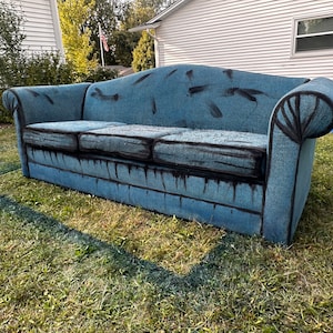 May include: A blue couch with black paint splatters on the fabric. The couch is sitting on a grassy lawn in front of a house.