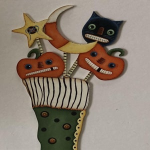 May include: Wooden Halloween decoration shaped like a stocking. The stocking is green with brown polka dots and a black heel. The top has a black and white striped pattern. The stocking is topped with a star, moon, cat, and two jack-o'-lanterns.