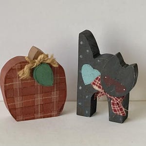 May include: Two wooden fall-themed decorations. One is a pumpkin with a brown and red plaid pattern, a green leaf, and a twine stem. The other is a black cat with a blue heart, a plaid scarf, and white polka dots.
