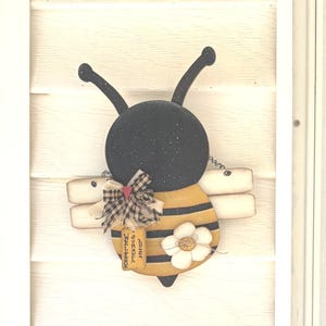 May include: A bee-shaped wall hanging featuring a black head with antennae, a yellow and black striped body, and white wings. It is decorated with a plaid fabric bow, a small red heart, a white flower, and a tag with the text "BEE HAPPY WELCOME".