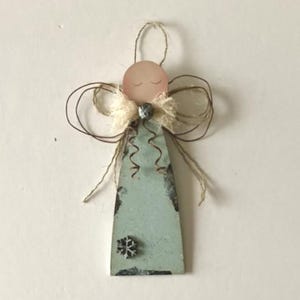 May include: A handcrafted angel ornament with a light blue body and a pink head. The angel features a snowflake detail, wire wings, and a jute string for hanging. The ornament is a rustic, decorative item.