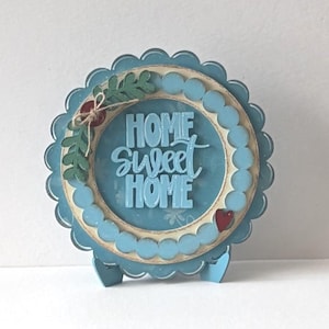 May include: A decorative, round, blue and cream-colored sign with the words "HOME Sweet HOME" in a script font. The sign has a scalloped edge and is supported by a stand. A sprig of greenery and a red heart add detail.