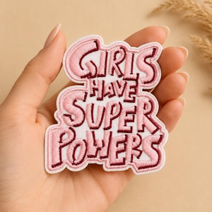 Könnte beinhalten: Gestickter Aufnäher in Rosa und Weiß mit dem Text "GIRLS HAVE SUPER POWERS". Der Aufnäher hat einen rosa Rand und wird in einer Hand gehalten. Der Hintergrund ist neutral. Ein lustiges Accessoire.