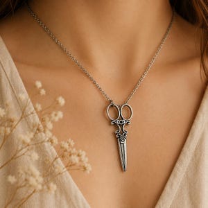 Può includere: Collana con pendente a forbice argentata. Il pendente presenta un design a forbice dettagliato con abbellimenti ornati. La collana è indossata su una delicata catena. Lo sfondo è di un colore neutro.