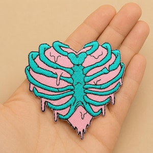 Op de afbeelding: Geborduurde patch in de vorm van een hart met een roze achtergrond en een turquoise ribbenkastontwerp. De patch heeft een druipend effect aan de onderkant. De patch wordt in een hand gehouden.