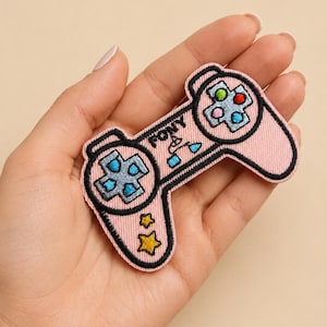 Peut inclure: Écusson rose brodé en forme de manette de jeu vidéo. La manette a des contours noirs et comporte des boutons bleus, rouges et verts. Le mot "PONY" est brodé sur la manette.