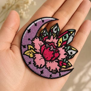 Op de afbeelding: Geborduurde patch met een paarse maan met zwarte sterren en een gedetailleerd bloemmotief. De bloem heeft roze en rode bloemblaadjes met gele en groene accenten, allemaal omlijnd in zwart.